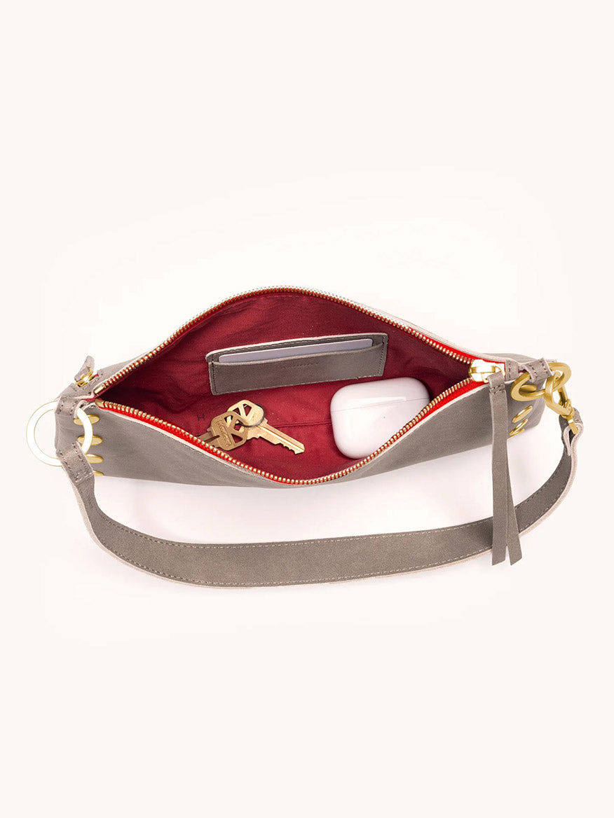 The Hammitt Los Angeles Kyle Shoulder Bag in Pewter/Brushed Gold/Red Zip is unzipped, revealing a set of keys, a small wallet, and white wireless earbuds nestled inside its red interior. This stylish convertible shoulder bag effortlessly transitions for any occasion.