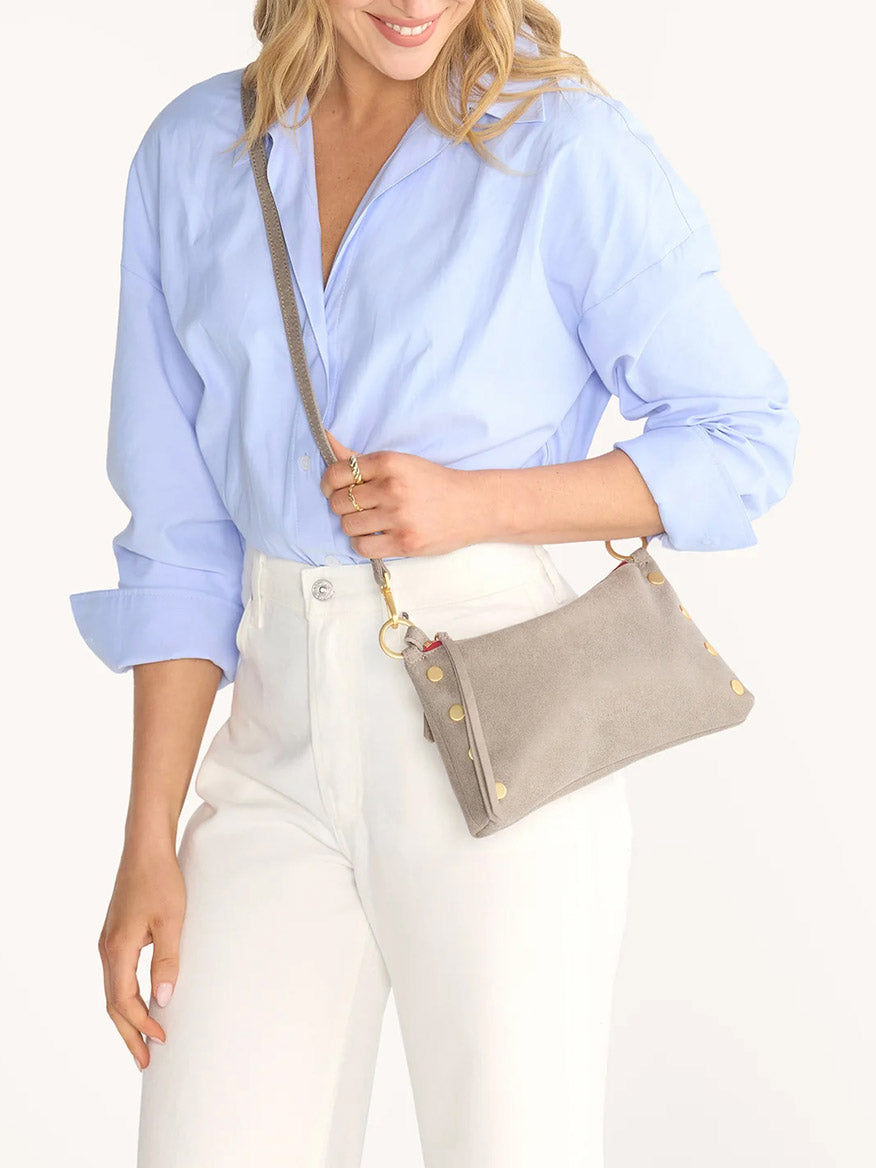 A woman wearing a light blue blouse and white pants holds the Hammitt Los Angeles Kyle Shoulder Bag in Pewter with brushed gold accents and a red zip against a plain background.