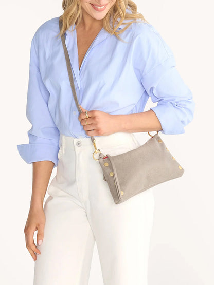 A woman wearing a light blue blouse and white pants holds the Hammitt Los Angeles Kyle Shoulder Bag in Pewter with brushed gold accents and a red zip against a plain background.