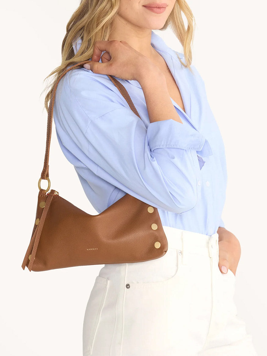 A woman in a light blue shirt and white pants holds the Hammitt Los Angeles Kyle Shoulder Bag in Mahogany Pebble with a red zip and gold hardware.