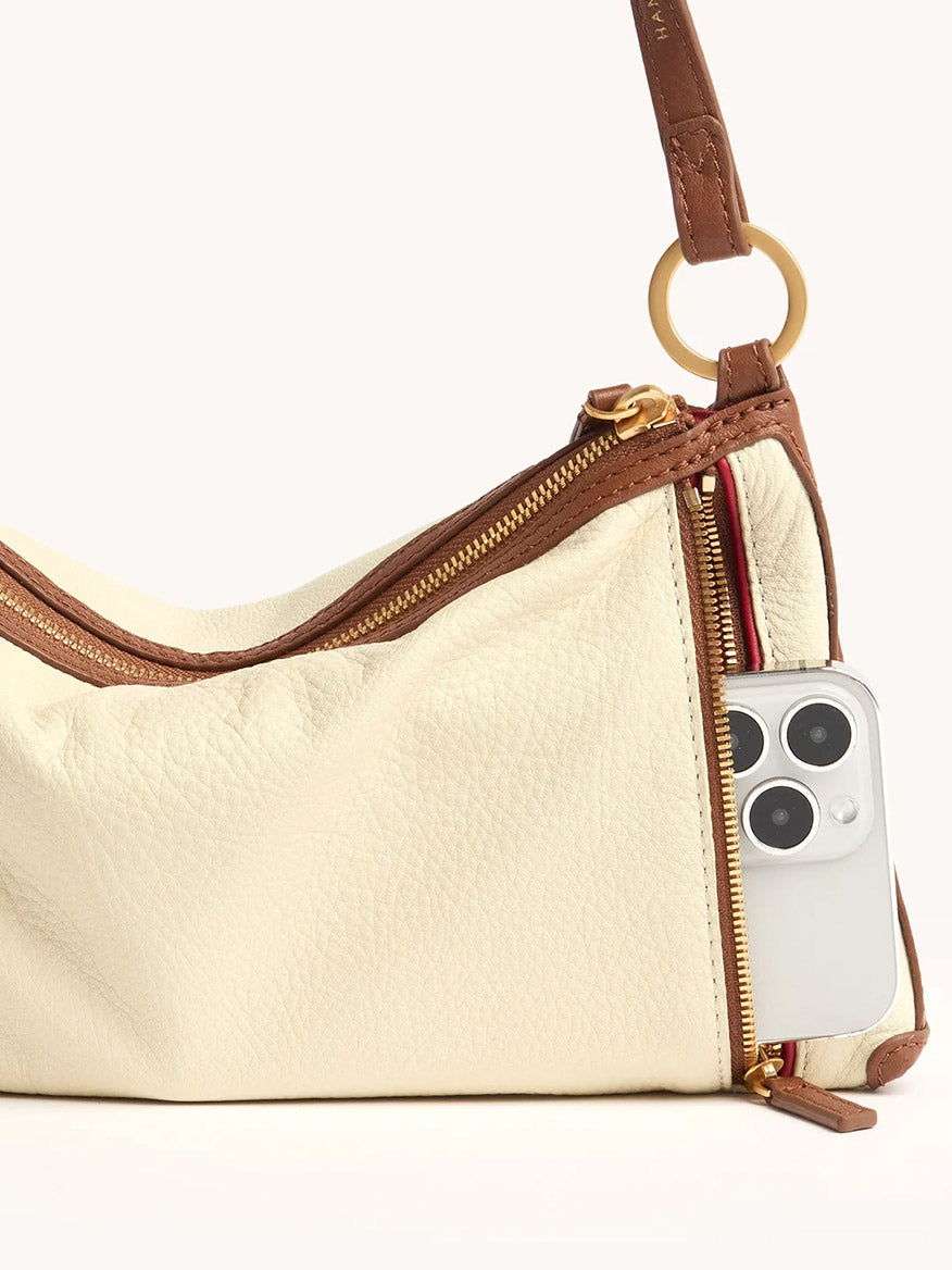The Hammitt Los Angeles Kyle Shoulder Bag in Courtyard Cream features pebbled cream leather with brown trim and a gold zipper, partially unzipped to reveal a silver smartphone in the outer pocket, plus a removable crossbody strap for versatility.