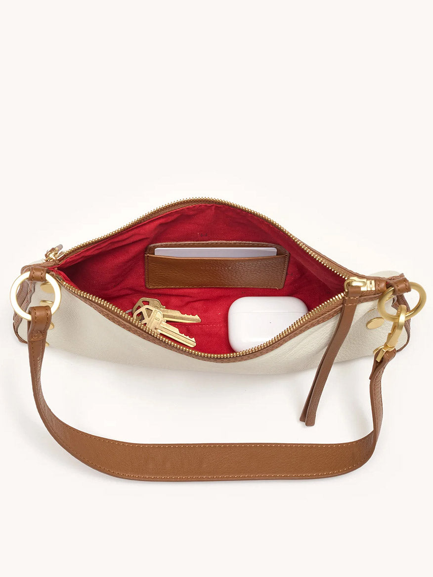 The Hammitt Los Angeles Kyle Shoulder Bag in Courtyard Cream, with its compact size and red interior, fits a phone, a set of keys, and a white wireless earbud case.