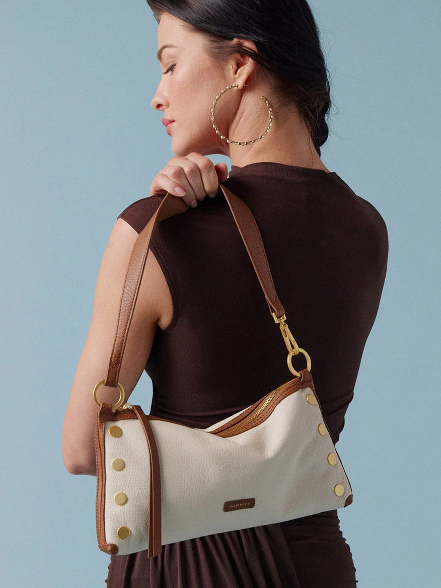 A woman in a brown dress holds the Hammitt Los Angeles Kyle Shoulder Bag in Courtyard Cream, featuring brown straps and gold hardware, over her shoulder while facing sideways against a light blue background.