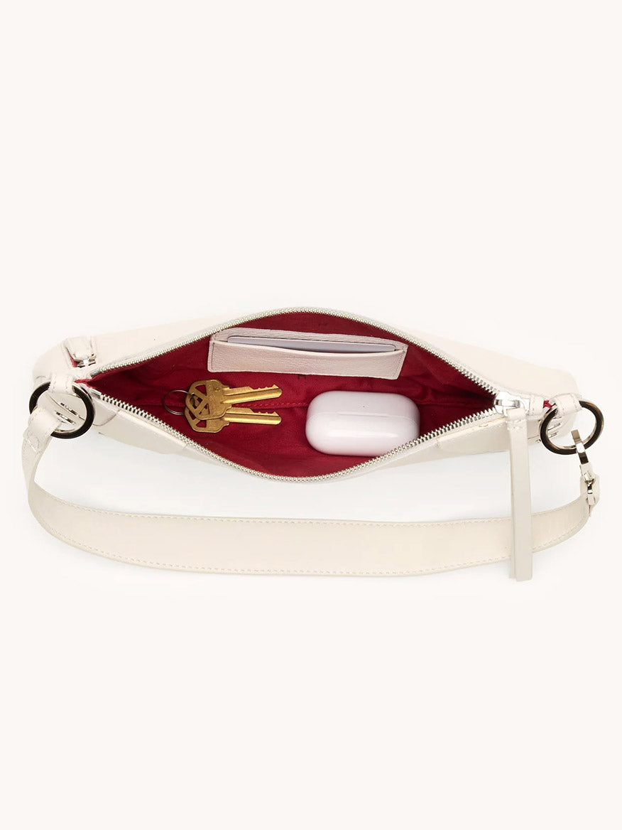 The Hammitt Los Angeles Kyle Shoulder Bag in Western Rein/Silver has white pebble leather, a red interior, and an adjustable crossbody strap. Shown top-down, it holds keys, a small wallet, and a white case.