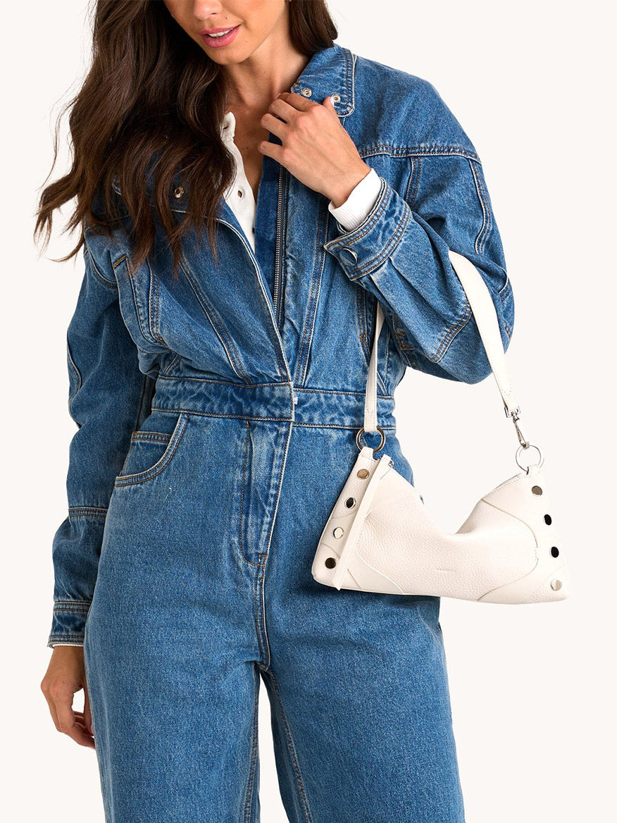 A woman in a blue denim jumpsuit holds the Hammitt Los Angeles Kyle Shoulder Bag in Western Rein/Silver, featuring metal rivets and an adjustable crossbody strap, standing against a plain background.