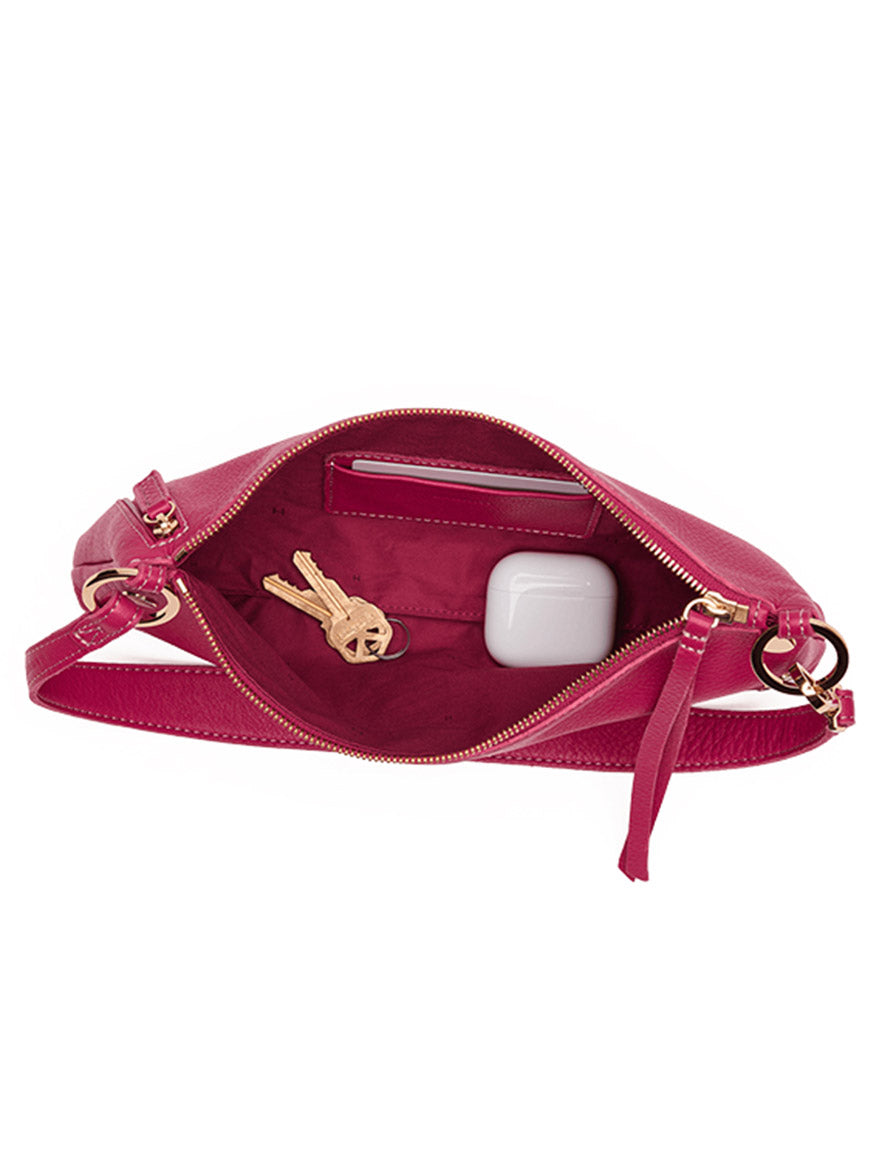 The Hammitt Los Angeles Kyle Shoulder Bag in Calypso Pebble/Gold, shown open from above, reveals a set of keys and a white wireless earbud case nestled inside its fuchsia pebbled leather interior and features a gold zipper closure.