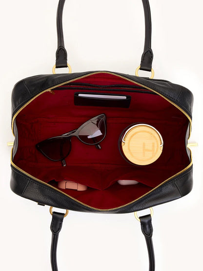 Top view of an open Hammitt Los Angeles Meredith Medium in Black/Brushed Gold, revealing its red interior with sunglasses, a wallet, a round container, and a small light-colored item inside.