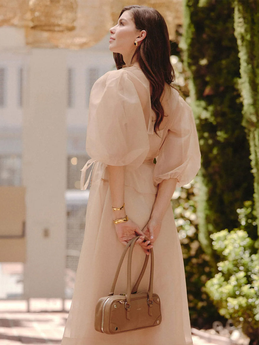 A woman outdoors in a light, sheer puff-sleeve dress holds the Hammitt Los Angeles Meredith Small in Dune/Silver—a chic doctor-style bag—behind her back, glancing to the side.
