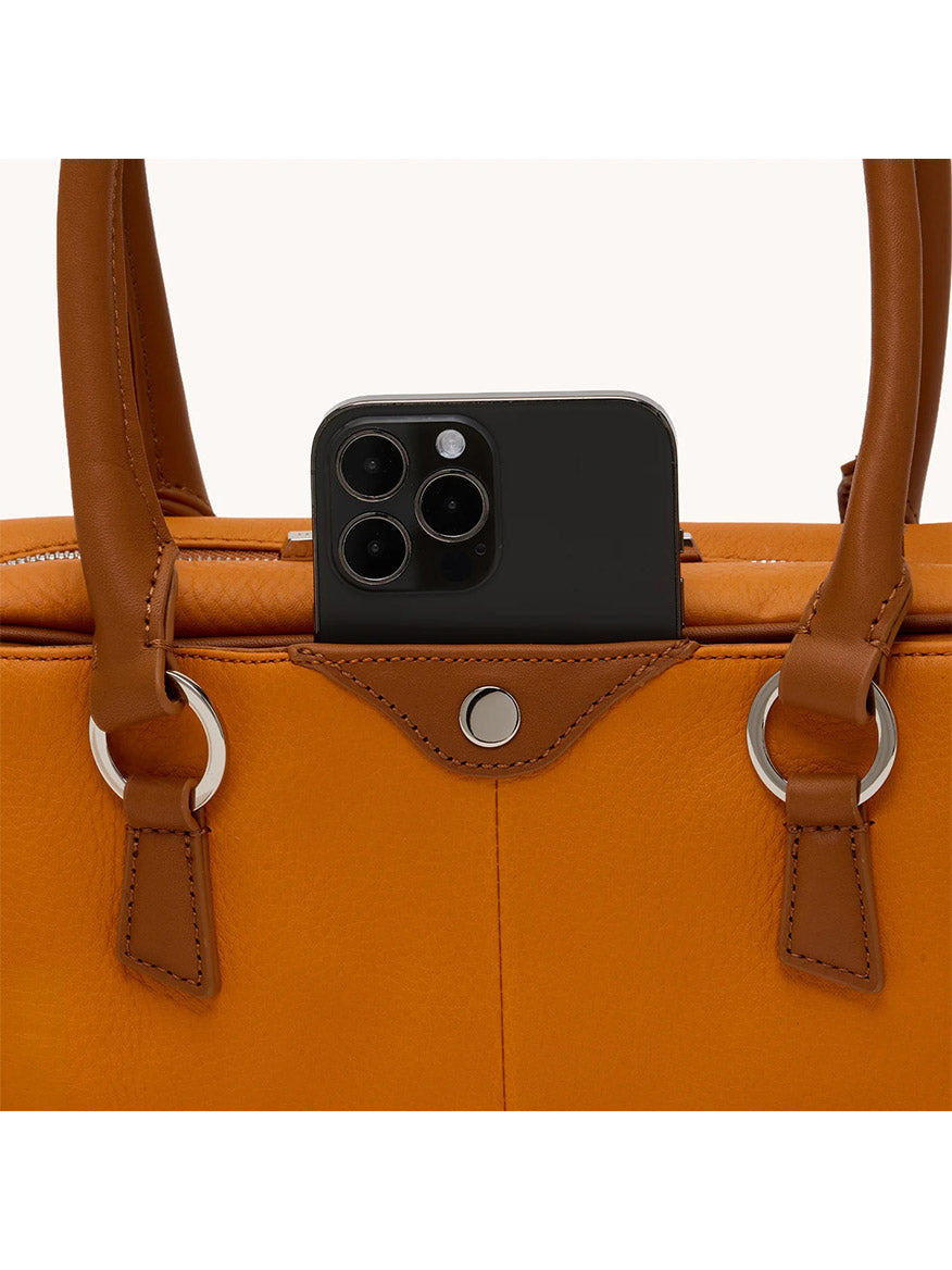 A black smartphone with three cameras is partially visible in the pocket of a Hammitt Los Angeles Meredith Small in Tailored Tangerine/Silver, an orange pebbled leather handbag with brown handles.