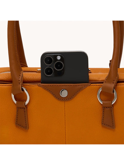 A black smartphone with three cameras is partially visible in the pocket of a Hammitt Los Angeles Meredith Small in Tailored Tangerine/Silver, an orange pebbled leather handbag with brown handles.