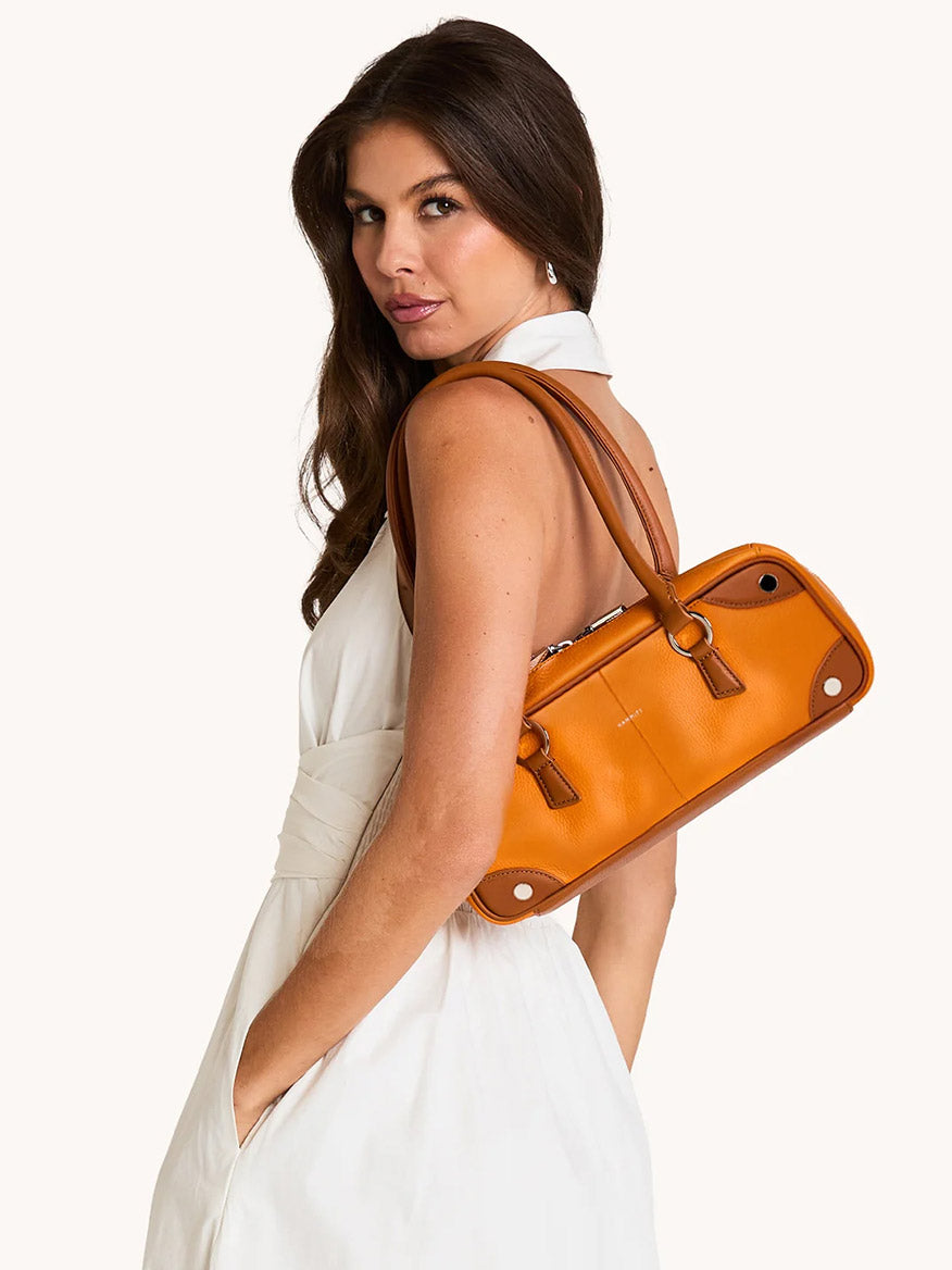 A woman in a sleeveless white dress poses with the Hammitt Los Angeles Meredith Small in Tailored Tangerine/Silver, glancing over her shoulder at the camera against a plain background.