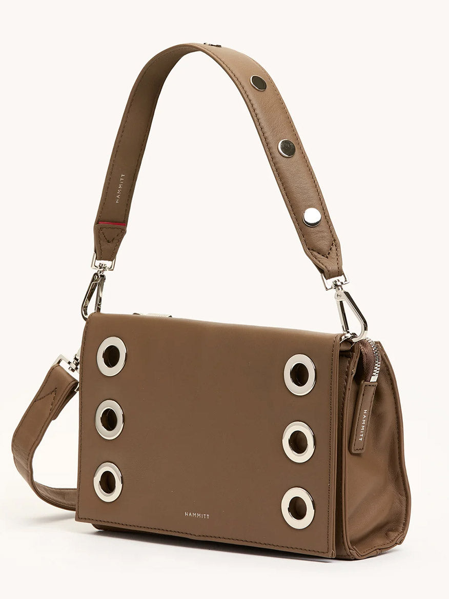The Hammitt Los Angeles Montana Clutch Sml in Dune/Silver is a rectangular taupe leather crossbody bag with six large silver grommets, a wide metal-detailed strap, and a zip closure—a chic, vintage-style accessory.