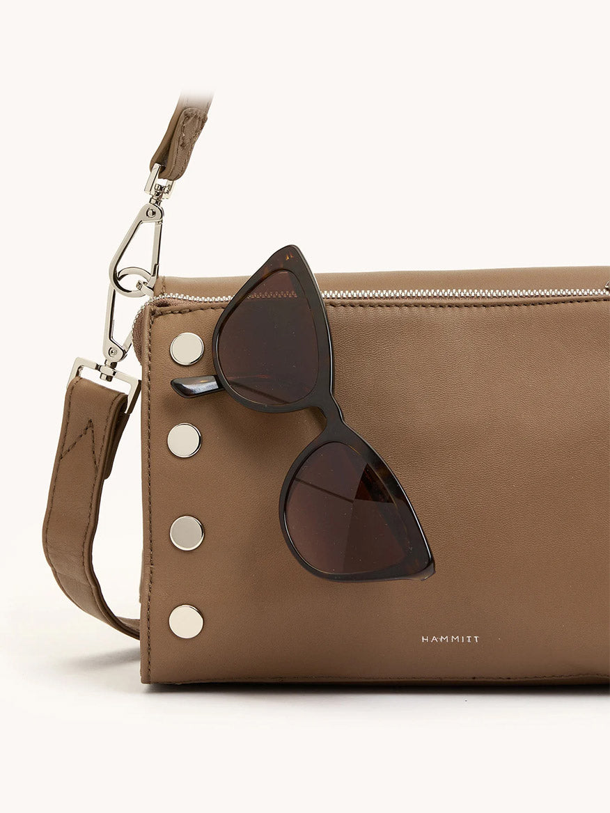 A pair of black cat-eye sunglasses rests on the Hammitt Los Angeles Montana Clutch Sml in Dune/Silver—a tan leather crossbody bag with silver studs and a detachable strap.