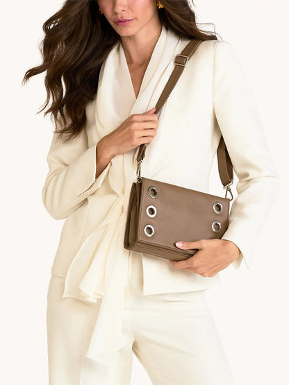 A woman in an off-white suit holds the Hammitt Los Angeles Montana Clutch Sml in Dune/Silver, a taupe vintage-style handbag featuring large silver grommets and a wide strap.