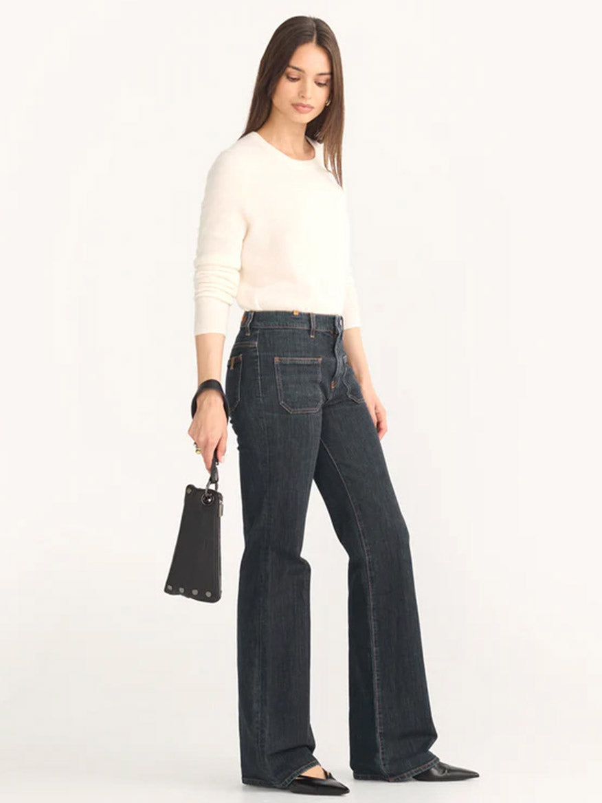 A woman in a white long-sleeve shirt and dark flared jeans is holding a Hammitt Los Angeles Nash Pochette in Black/Gunmetal, highlighting its sleek pebbled leather finish.