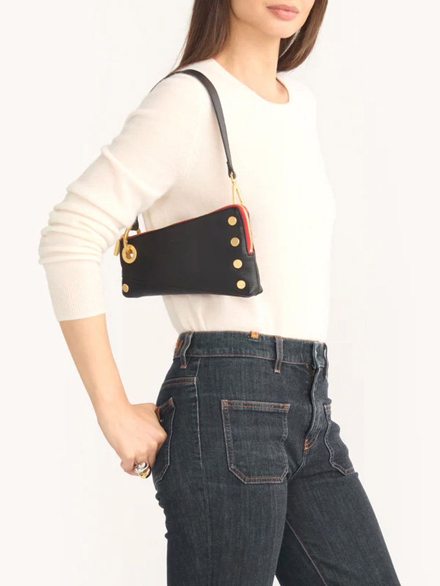 A person in a white sweater and dark jeans holds a stylish Hammitt Los Angeles Nash Pochette in Black/Brushed Gold & Red Zip on their shoulder. The black handbag, crafted from pebbled leather, features elegant gold accents and a detachable J-hook strap for added versatility.