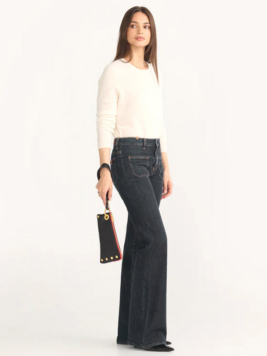 A woman stands wearing a white sweater and dark wide-leg jeans, holding a Hammitt Los Angeles Nash Pochette in Black/Brushed Gold with a Red Zip.