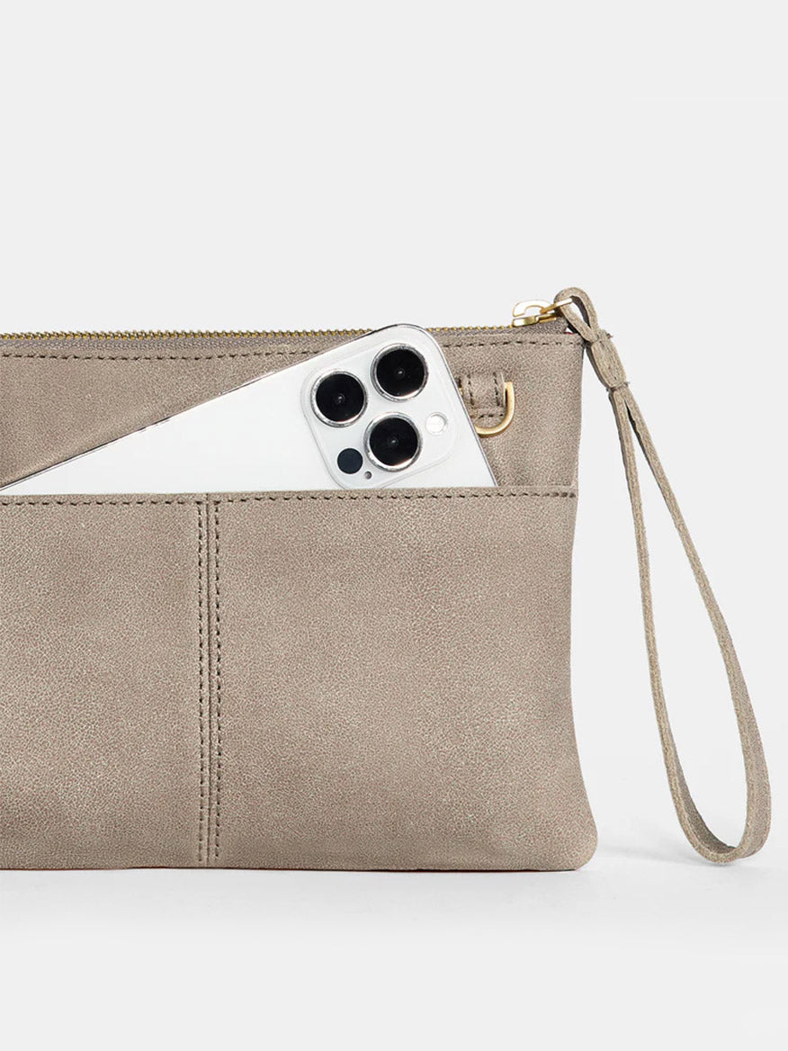 A Pewter Hammitt Los Angeles Nash Small Clutch with a zipper and wrist strap, partially open to reveal a white smartphone inside. The versatile clutch includes a convertible strap for easy transformation into a chic crossbody accessory.