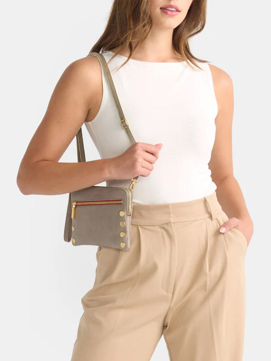 A person wearing a white sleeveless top and beige trousers stands holding the Hammitt Los Angeles Nash Small Clutch in Pewter, featuring gold buttons and a convertible strap.