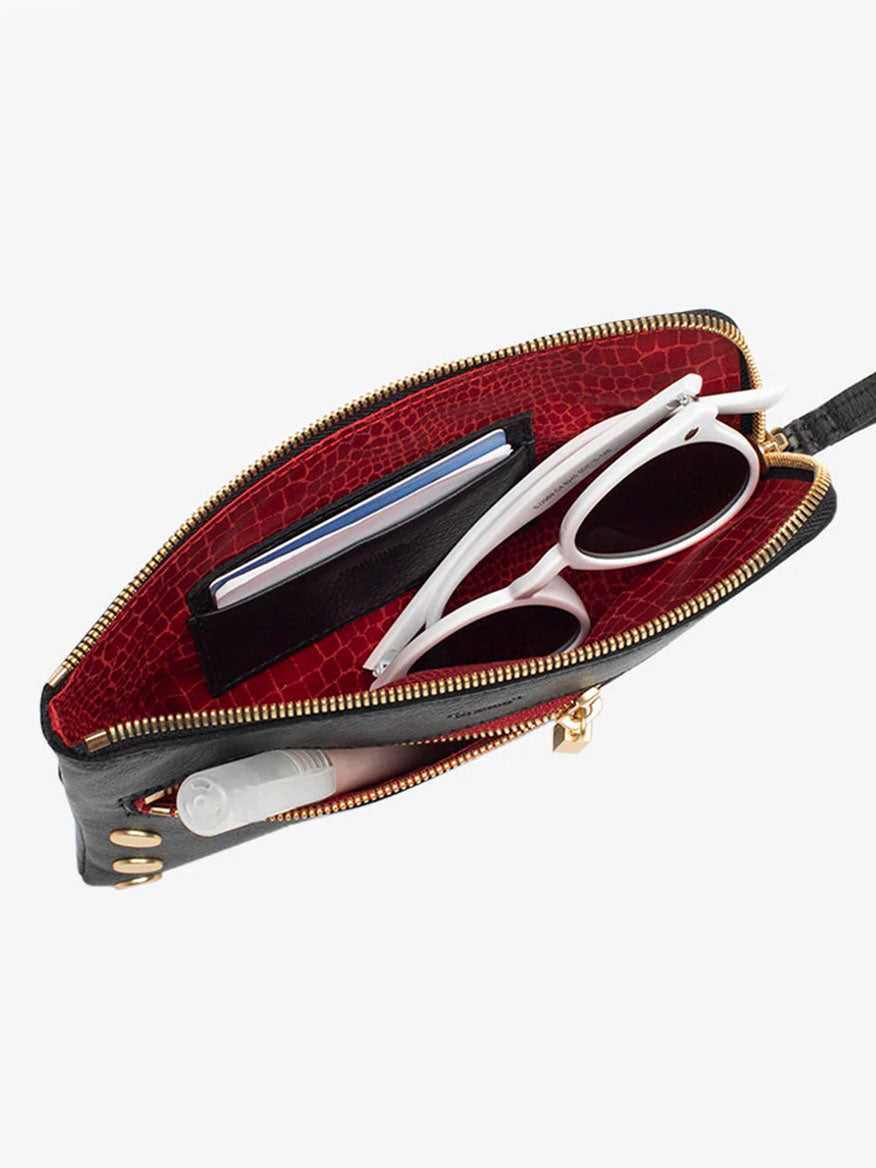 A top-view of an open Hammitt Los Angeles Nash Small Clutch in Black with Brushed Gold and Red Zip reveals a luxurious red interior holding a white pair of sunglasses, a black wallet, and other small items. The clutch is designed for added versatility with its removable crossbody strap.