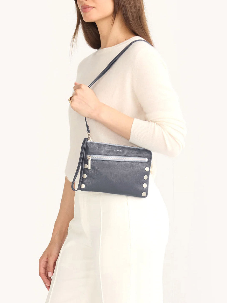 A person wearing a cream sweater and white pants holds the Hammitt Los Angeles Nash Small Clutch in Jazz Blues, a navy blue crossbody bag crafted from pebbled leather with a zippered pocket and decorative buttons.