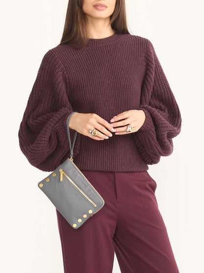 A person wearing a burgundy sweater and pants holds a Hammitt Los Angeles Nash Small Clutch in Sonnet Grey, featuring gold details and a convertible strap.