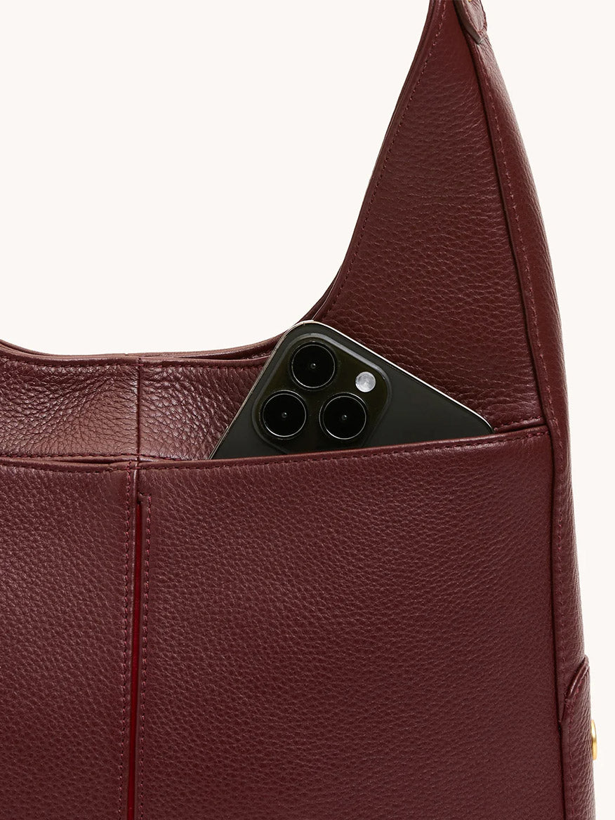 A black smartphone peeks out from the outer pocket of the Hammitt Los Angeles Tim Med Hobo in Cherrywood Pebble/Brushed Gold.