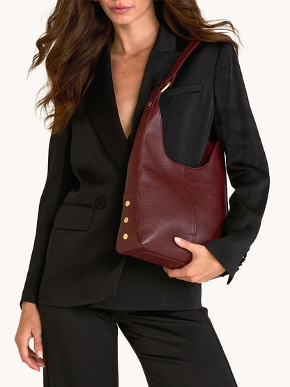 A woman in a black suit holds the Hammitt Los Angeles Tim Med Hobo in Cherrywood Pebble/Brushed Gold, featuring gold rivet details, against a plain background.