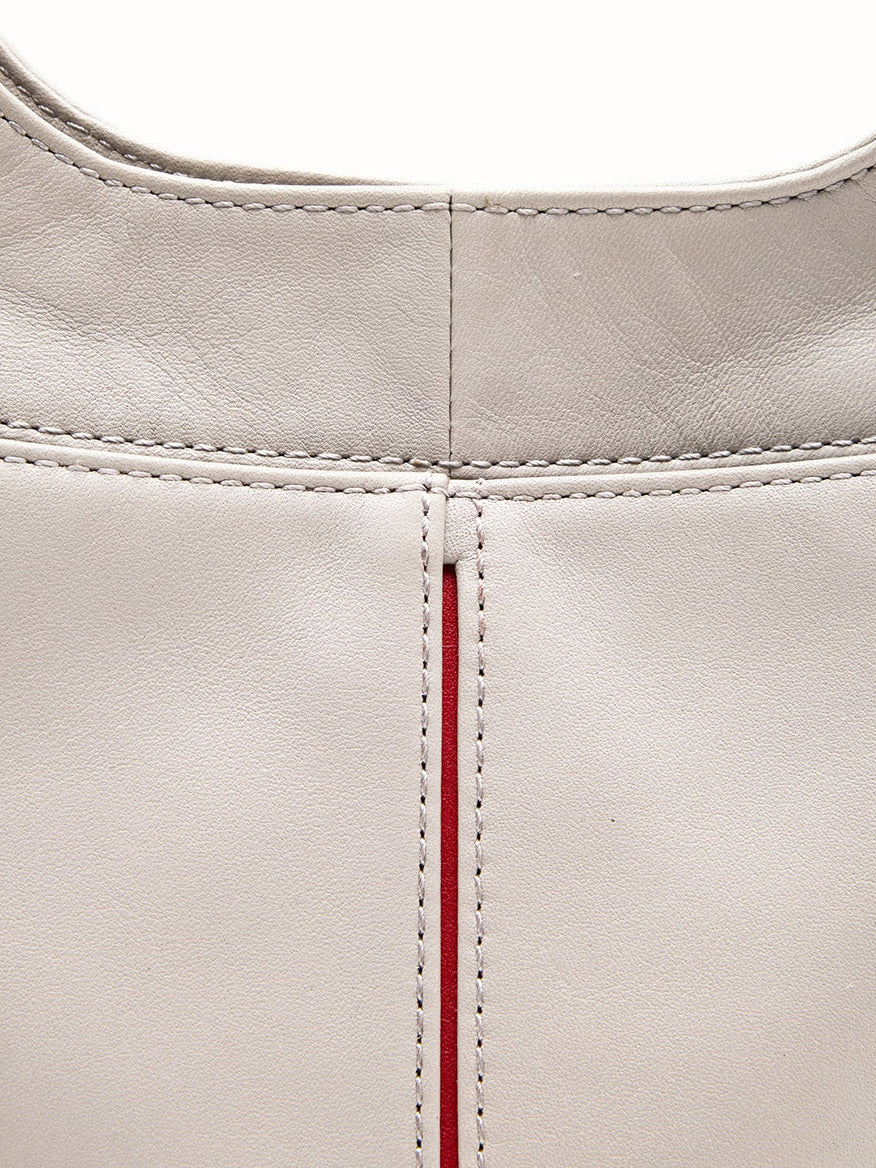 Close-up view of the Hammitt Los Angeles Tim Med Hobo in Stormy/Brushed Gold, showcasing its light gray pebble leather surface with visible stitching and a vertical seam accented by a thin red stripe.