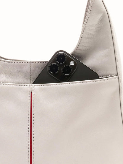 A smartphone peeks from the outer pocket of the Hammitt Los Angeles Tim Med Hobo in Stormy/Brushed Gold, featuring a light gray hue and red stitched accents.