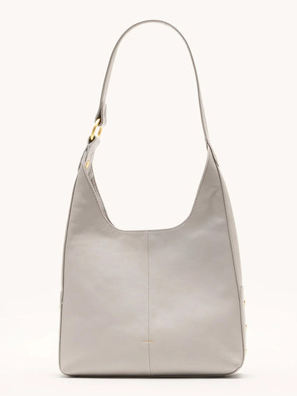 The Hammitt Los Angeles Tim Med Hobo in Stormy/Brushed Gold is a minimalist, light gray pebble leather bag with a wide strap and gold-tone hardware, displayed upright against a plain background.