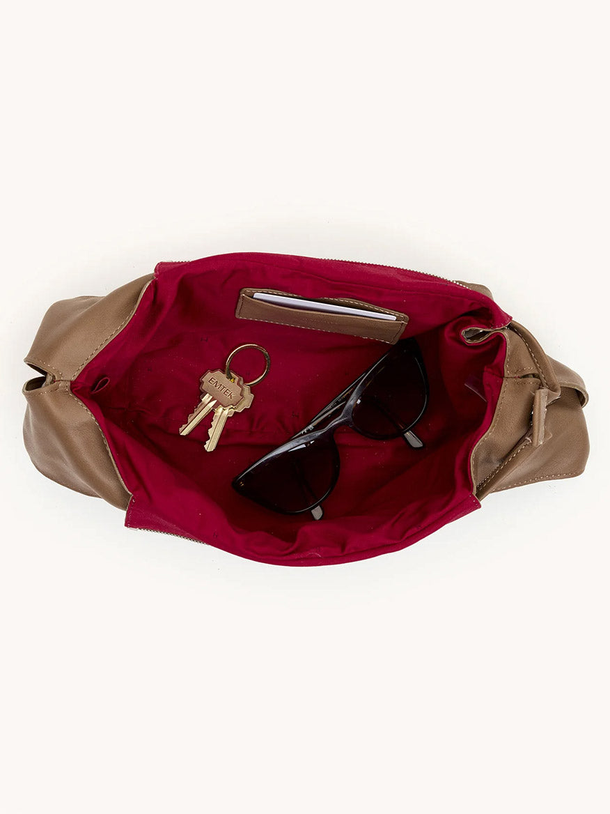 The Hammitt Los Angeles Tom Zip Medium in Dune/Silver is an open tan hobo bag with red lining and an adjustable shoulder strap. Inside its zippered compartment are keys, black sunglasses, and a slim silver phone.