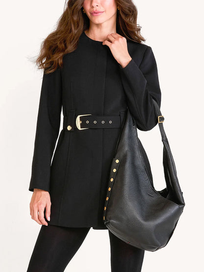 Woman wearing a long-sleeve black belted blazer holds the Hammitt Los Angeles Tom Zip Large in Black/Brushed Gold/Red Zip, a spacious bag with gold accents, standing against a plain light background.