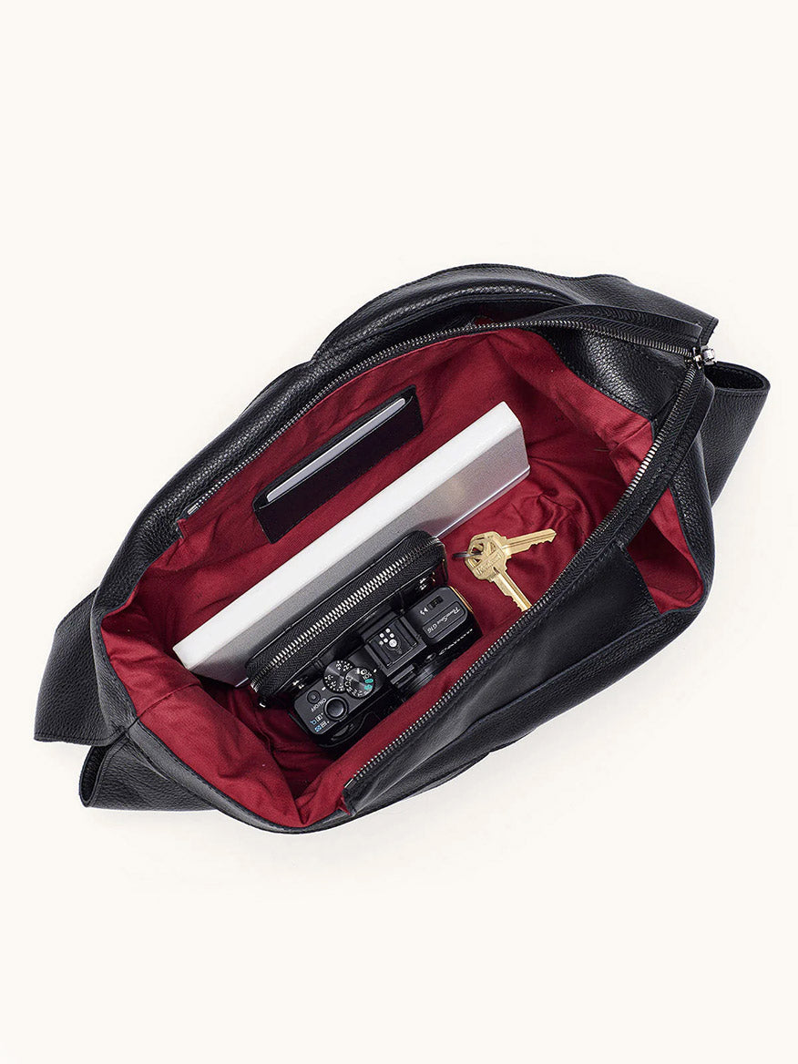 Top view of an open Hammitt Los Angeles Tom Zip Large in Black/Gunmetal, with a red lining, holding a white rectangular object, black case, set of keys, and a black camera.