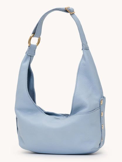 The Hammitt Los Angeles Tom Zip Medium in Hazy Blue/Gold is a light blue pebbled leather compact hobo bag with a wide adjustable shoulder strap, gold buckle, and decorative gold buttons along the side.