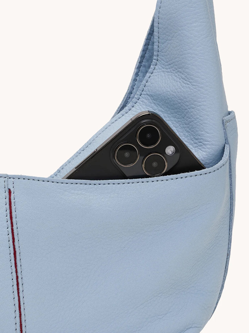 A black smartphone is partially visible in the outer pocket of the Hammitt Los Angeles Tom Zip Medium bag in Hazy Blue/Gold, crafted from pebbled leather.