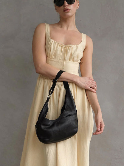 A woman in a sleeveless, cream dress and black sunglasses holds the Hammitt Los Angeles Tom Zip Medium in Black/Gunmetal, a pebbled leather hobo bag with an adjustable shoulder strap, against a plain gray background.