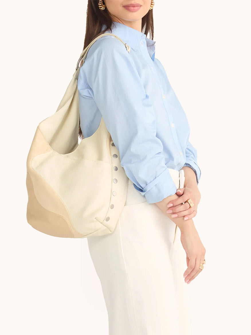 A person in a light blue shirt and white pants is holding the Hammitt Los Angeles Tom Zip in Coastal Cream with an adjustable strap on their shoulder.