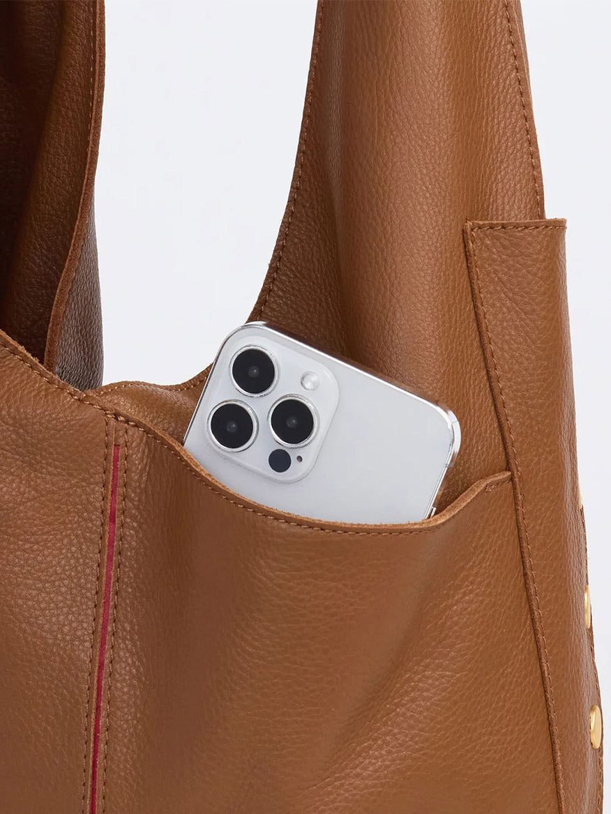 A smartphone with a white case peeks from the pocket of the Hammitt Los Angeles Tom Zip in Mahogany Pebble bag, highlighting its elegant design and adjustable strap.