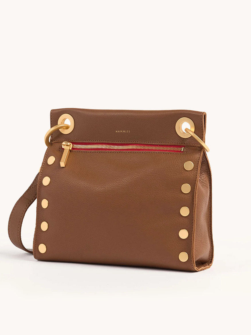 The Hammitt Los Angeles Tony Medium Crossbody Bag in Mahogany Pebble w/Red Zip features gold circular studs, a front red zippered pocket, and large gold rings near the top corners. Crafted from pebbled leather, it includes an adjustable crossbody strap for versatile styling.