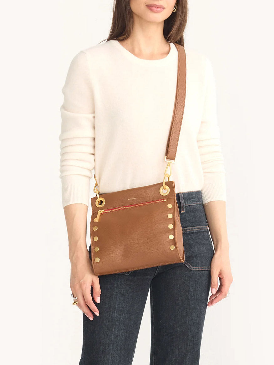 A person in a white sweater and jeans holds the Hammitt Los Angeles Tony Medium crossbody bag in mahogany pebble with a red zip, featuring gold studs and an adjustable strap.