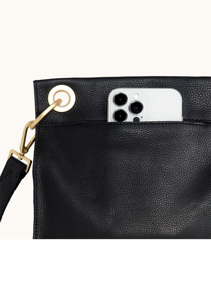 A Hammitt Los Angeles Tony Medium Crossbody Bag in Black/Brushed Gold/Red Zip with an outer pocket holds a silver smartphone featuring three camera lenses.