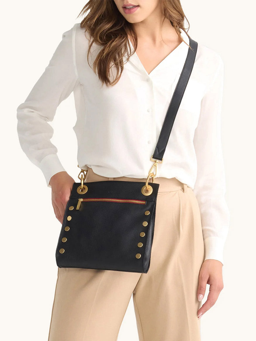 Woman in a white blouse and beige pants carries the Hammitt Los Angeles Tony Medium Crossbody Bag in Black/Brushed Gold/Red Zip—an ideal everyday accessory with gold accents and a front zipper pocket.