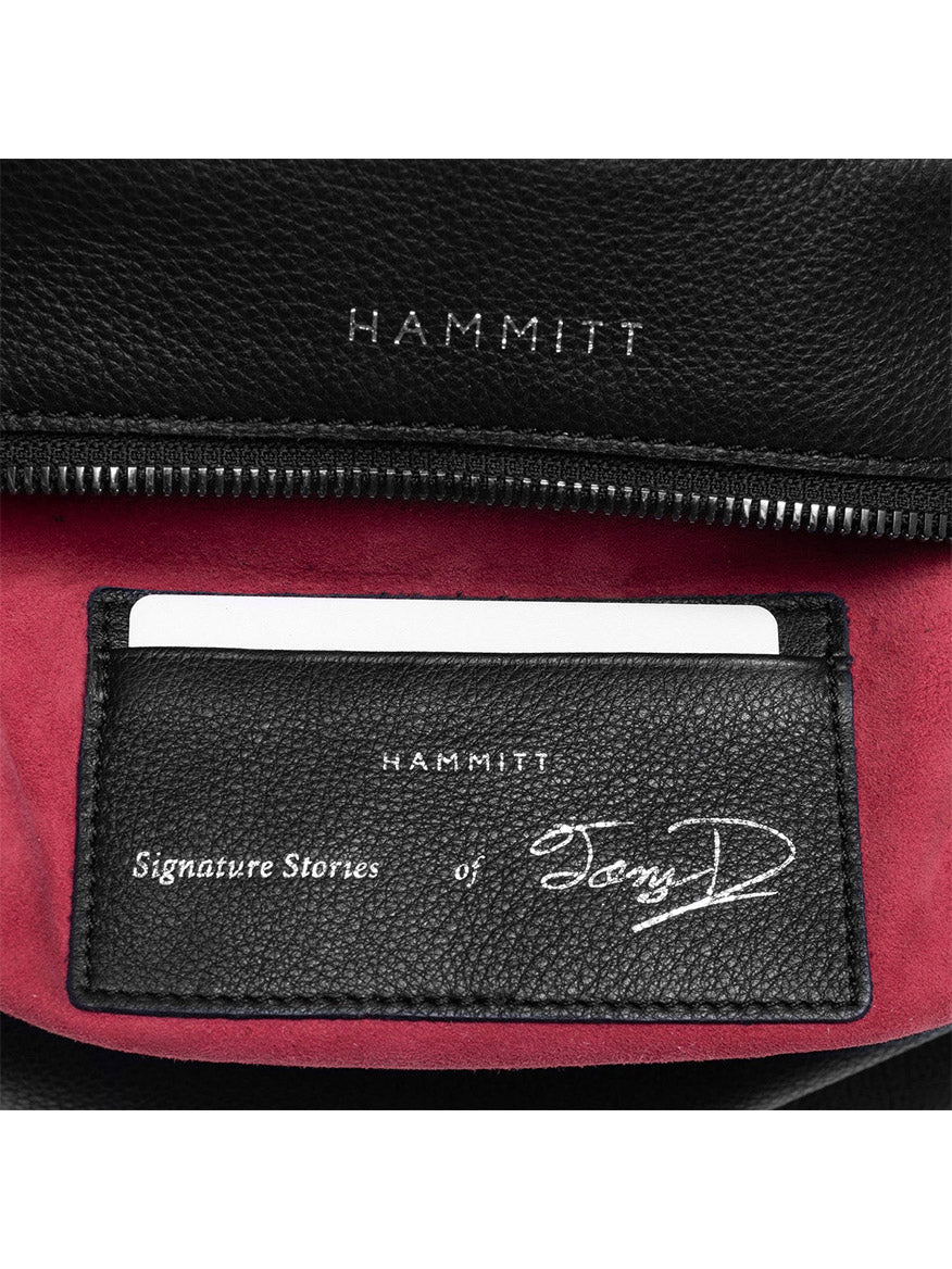 Close-up of the Hammitt Los Angeles Tony Signature Medium Crossbody in Black & Gunmetal, highlighting black pebbled leather with red lining and an interior label "Signature Stories of Tony D" alongside a credit card slot. This stylish crossbody offers an adjustable strap for versatile use.