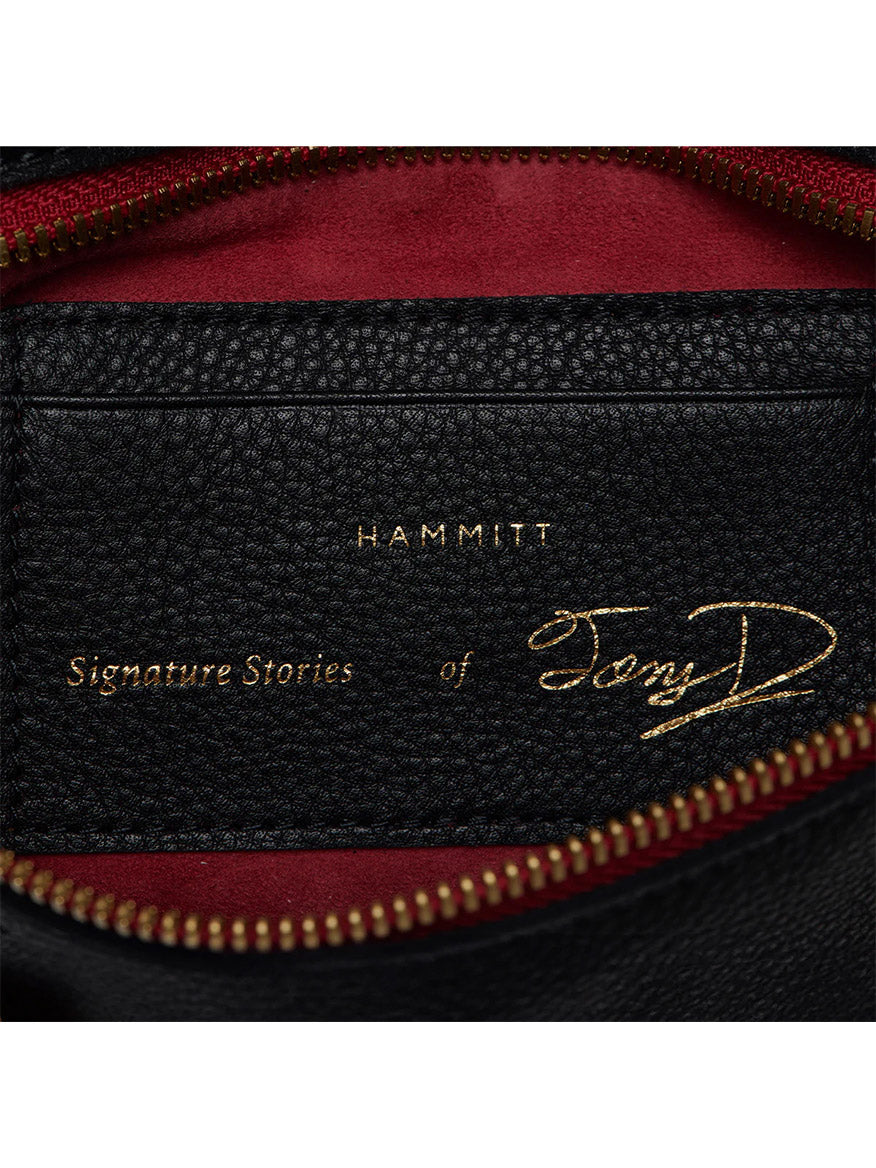 Close-up of a black leather Hammitt Los Angeles Tony Signature Medium Crossbody in Black with text reading "HAMMITT Signature Stories of Tony D" in gold lettering, shown against a background of a partially unzipped red fabric lining and adorned with limited edition commemorative details.