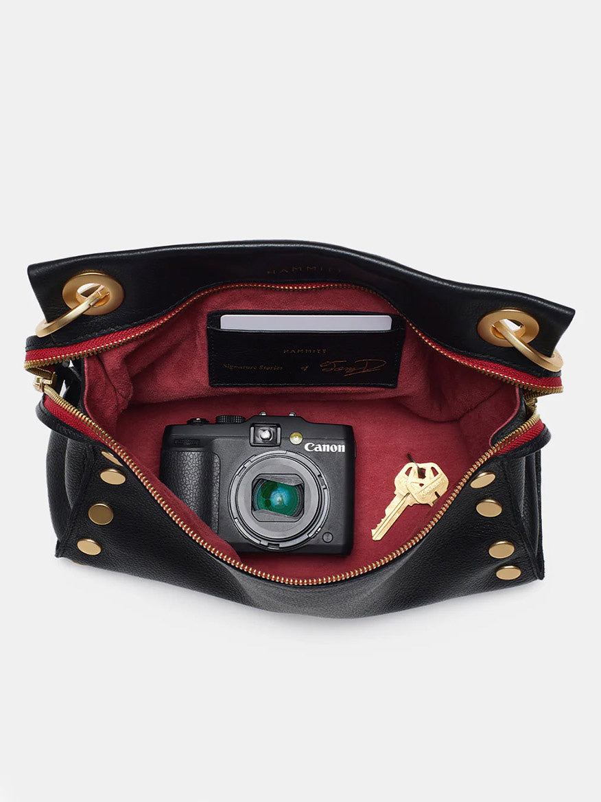 An open Hammitt Los Angeles Tony Signature Medium Crossbody in Black with red interior lining reveals a limited edition Canon camera, a brass key, and a closed passport inside.