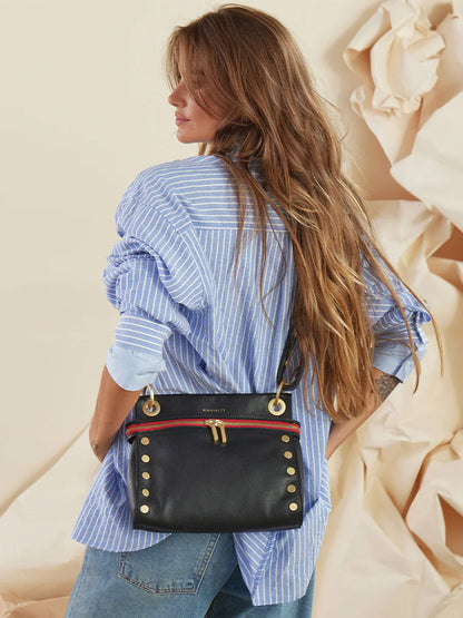 A person with long hair wearing a blue striped shirt and jeans is carrying a Hammitt Los Angeles Tony Signature Medium Crossbody in Black over their shoulder. The background features crumpled beige paper.