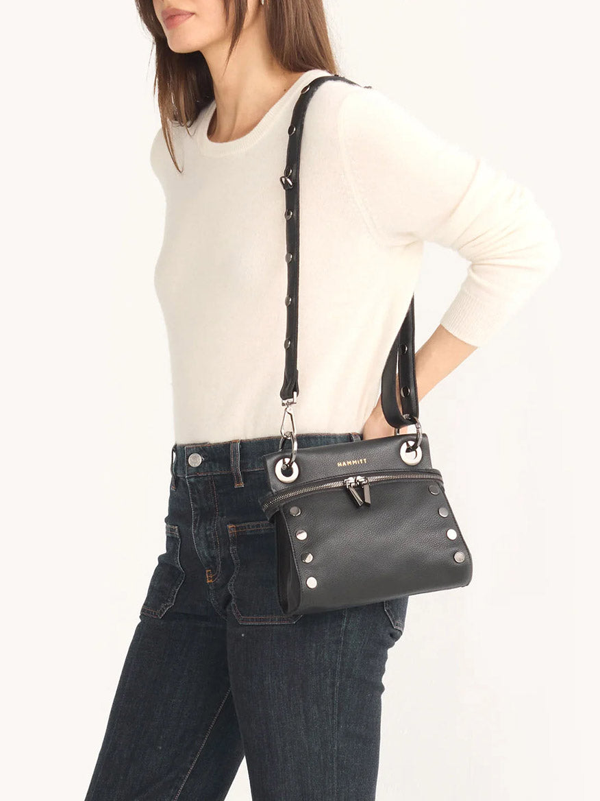 A person wearing a white sweater and jeans carries the Hammitt Los Angeles Tony Signature Small Crossbody in Black, crafted from pebbled leather with silver studs on its adjustable strap.