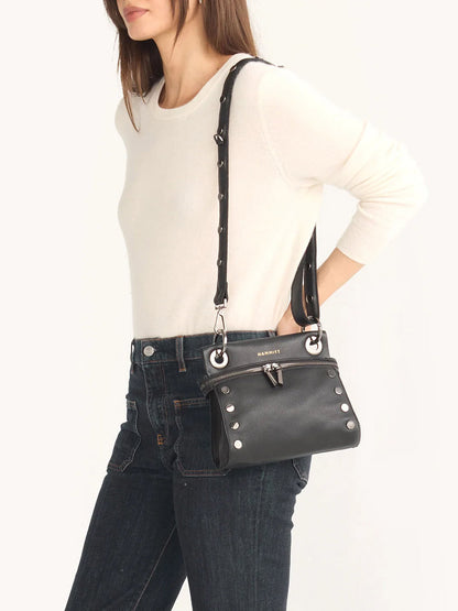 A person wearing a white sweater and jeans carries the Hammitt Los Angeles Tony Signature Small Crossbody in Black, crafted from pebbled leather with silver studs on its adjustable strap.