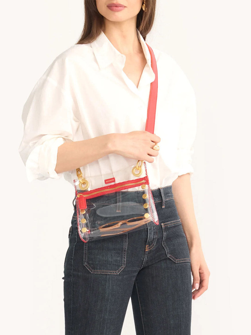 Wearing a white shirt and jeans, a person confidently holds the Hammitt Los Angeles Tony Small Crossbody Bag in Clear Poppy Red, which features an adjustable vibrant red strap. This stylish yet functional accessory is stadium-approved, ensuring you breeze through security effortlessly.